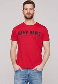 Red cotton t-shirt with short sleeves and a crew neck. Features bold black text "CAMP DAVID" and a casual fit, paired with light blue jeans.