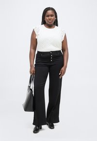 Γυναίκα με πλεξούδες στα μαλλιά, φοράει λευκό αμάνικο τοπ με διακοσμητικές τρύπες, μαύρο παντελόνι φαρδύ στο πόδι με ψηλή μέση, μαύρες μποτάκια μέχρι τον αστράγαλο, κρατώντας μαύρη τσάντα tote.