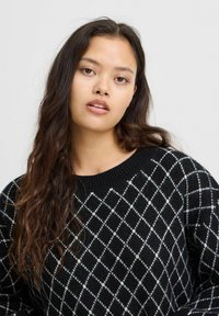 Pull en tricot noir avec un motif losange en blanc. Le design présente un col rond et des poignets côtelés. Texture douce, coupe décontractée.