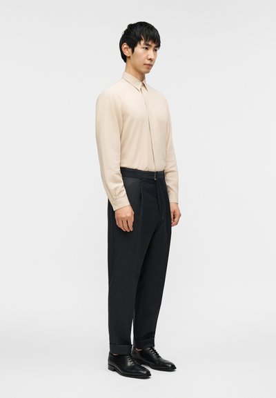 Άνδρας όρθιος που φοράει μπεζ πουκάμισο με μακριά μανίκια, σκούρο παντελόνι με ριγέ ψηλή μέση και μανσέτα, και μαύρα παπούτσια φόρμας σε λευκό φόντο.