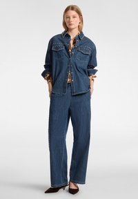 Giacca di jeans con colletto borchiato e tasche, abbinata a pantaloni in denim a gamba larga. Entrambi in una tonalità blu medio con una texture liscia.