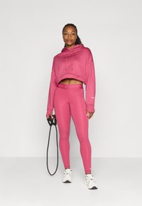 Cropped mikina a legíny Nike Pro v žiarivej ružovej farbe, s logom na hrudi a vysokým pásom. Modelka drží švihadlo.