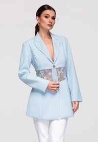 Mujer con blazer azul claro con detalle de encaje en la parte media y botón dorado, combinada con pantalones blancos, posando contra un fondo liso.