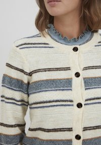 Svītrots kardigāns krēmkrāsā, zilā, brūnā un oranžā krāsā. Tam ir apaļa apkakle ar volānu maliņu, pogu aizdare un tekstūrēta adījuma. 