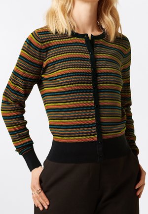 Frau trägt eine taillierte, geknöpfte Strickjacke mit horizontalen, strukturierten Streifen in Grün, Blau, Pink, Senf und Schwarz, kombiniert mit schwarzen Hosen.