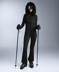 Czarny jednoczęściowy kombinezon narciarski z teksturowanym górnym częściami, kapturem wyłożonym futrem i rozszerzanymi nogawkami. Noszony z lustrzanymi goglami i kijami narciarskimi. Czarne buty narciarskie.