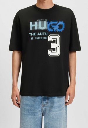 Mężczyzna w czarnej koszulce z niebiesko-białym tekstem "HUGO", "THE AUTU", "LIMITED TICKE" oraz dużą cyfrą 3, w parze z jasnoniebieskimi dżinsami.
