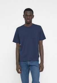 Μπλε ναυτικό βαμβακερό μπλουζάκι με στρογγυλή λαιμόκοψη, κοντά μανίκια και τσέπη στο στήθος. Διαθέτει μια μικρή ετικέτα μάρκας στη ραφή πλευράς.