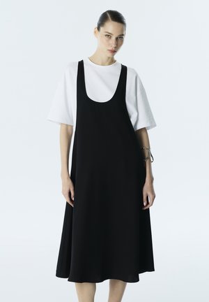 Kobieta z zaczesanymi do tyłu włosami, ubrana w luźną czarną sukienkę bez rękawów na białym, oversizeowym t-shircie, stojąca na tle jednolitego jasnego tła.