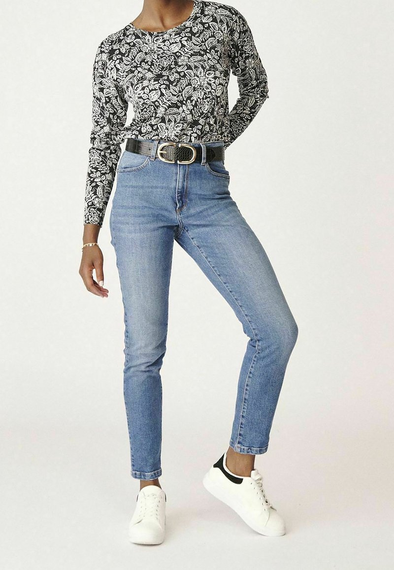 Personne portant un haut à manches longues à motif floral noir et blanc, un jean taille haute bleu clair avec une ceinture noire, et des baskets blanches avec des accents noirs.