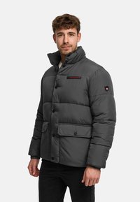 Man draagt een donkergrijze donzen jas met hoge kraag, knopen aan de voorkant, klepzakken en een rood "Kronstadt" label op de borst.