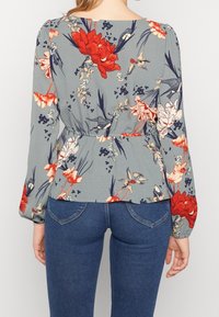 Blumenbluse aus hellgrauem Stoff, verziert mit roten und pinken Blumen sowie grünen Akzenten, elastischem Bund und langen Ärmeln. Kombiniert mit blauen Jeans.