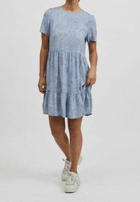 Kvinna i en kortärmad, knälång blå klänning med vita prickar och vita snörskor som står mot en vit bakgrund.