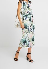 Jumpsuit w kwiatowy wzór o luźnym kroju, z krótkimi rękawami zdobionymi koronką, z talią wciętą i długością do połowy łydki. W połączeniu z czarnymi sandałami na słupku.