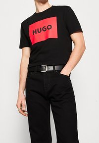 T-shirt in cotone nero con un grande rettangolo rosso che riporta la scritta "HUGO" in nero. Indossato con pantaloni neri e una cintura nera.