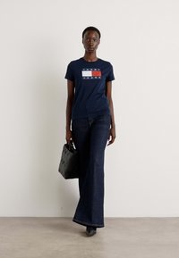 Námornícka modrá grafická tričko s textom "TOMMY JEANS", kombinované s tmavými džínsovými zavinovacími nohavicami a čiernou vzorovanou kabelkou. Hladké textúry.