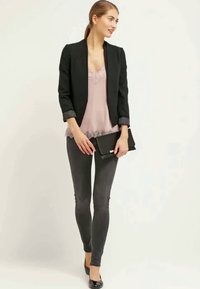 Mulher com blazer preto, blusa rosa com renda, jeans skinny escuros, sapatos de salto pretos, segurando uma clutch preta, olhando para o lado em frente a um fundo simples.