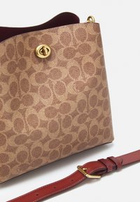 Tan coated fabric bag with a patterned design, featuring a gold snap closure and a red leather adjustable strap.
