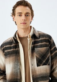 Camisa de franela multicolor con patrones de cuadros en marrón, crema y negro. Diseñada con un bolsillo y cierre de botones, superpuesta sobre un suéter beige.