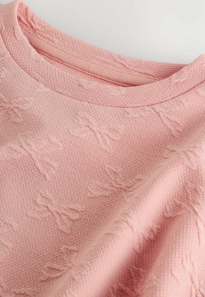 Jasnoróżowy sweter z fakturowanej tkaniny z wytłaczanymi wzorami w kształcie kokardy. Posiada klasyczny okrągły dekolt i miękką, tkana strukturę.