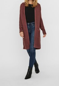 Długi, bordowy sweter z kapturem na drutach, z przednimi kieszeniami, prążkowanymi mankietami i dołem, w zestawieniu z ciemnymi dżinsami i botkami za kostkę.