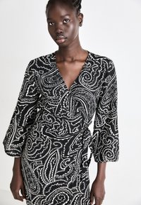 Vestito avvolgente paisley bianco e nero con maniche lunghe a campana, scollatura a V profonda e una cintura annodata in vita. Tessuto liscio e strutturato.