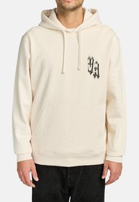 Man in een crèmekleurige hoodie met een buidelzak en een zwart gotische "VA" logo op de linkerkant van de borst, gecombineerd met zwarte broek.