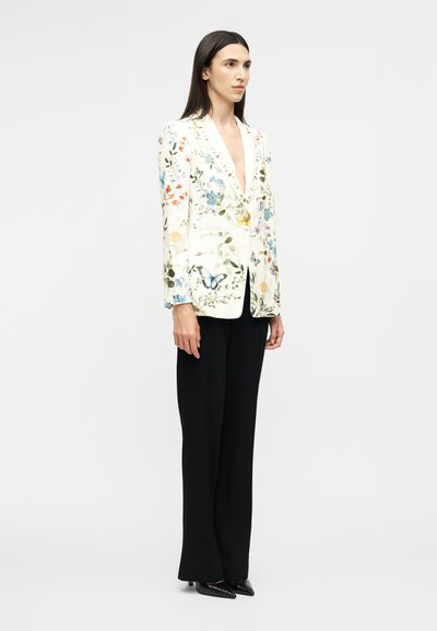 Γυναίκα με μακριά σκούρα μαλλιά που φοράει λευκό σακάκι με φλοράλ σχέδιο πεταλούδας, μαύρο παντελόνι φαρδύ και μαύρες μυτερές γόβες, όρθια.