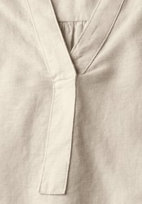 Camicia di lino color beige chiaro con scollatura a V, caratterizzata da un dettaglio di patta, tessuto finemente texturizzato e schiena leggermente arricciata per una vestibilità rilassata.