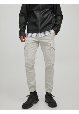 Personne portant un pantalon cargo beige avec des chevilles élastiques, des baskets noires, une veste en cuir noire et une chemise à carreaux en dessous.