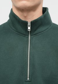 Tmavě zelená zipová mikina s vysokým límcem a stříbrným zipem. Vyrobená z měkké, texturované látky, s minimalistickým designem.