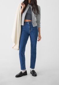 Jeans in denim blu a vita alta abbinati a un cardigan grigio, mocassini neri e un top grigio a coste; la modella tiene un maglione color crema.