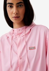 Rosa wasserdichte Kapuzenjacke mit Druckknöpfen, verstellbarem Kordelzug und einem kontrastierenden schwarz-roten Markenpatch auf der Brust.