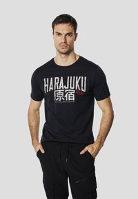 T-shirt en coton noir avec un graphisme audacieux « HARAJUKU » en blanc et rouge. Col rond, manches courtes, texture lisse, porté avec un pantalon noir.