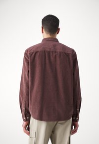 Camisa de pana burdeos con cuello abotonado, mangas largas y puños abotonados. Textura suave y dobladillo ligeramente curvado en la parte posterior.