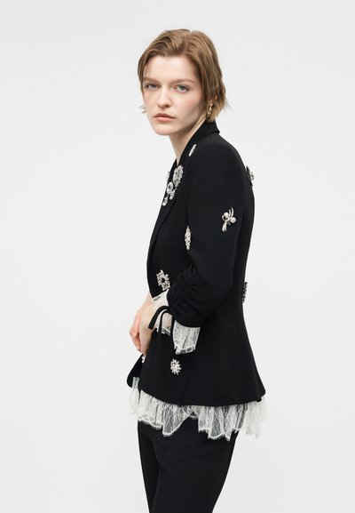 Νεαρή γυναίκα με κοντά μαλλιά, φορεί μαύρο σακάκι διακοσμημένο με ασημένια καρφίτσες και άσπρες δαντελένιες μανσέτες και τελείωμα, πάνω από μαύρο παντελόνι.