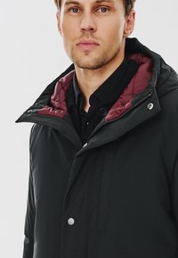 Czarna pikowana kurtka z bordowym podbiciem, z zapięciem na zamek, zatrzaskami i kapturem. Gładka tekstura o schludnym designie.