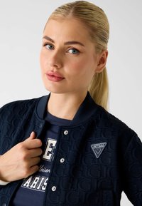 Marineblaue Bomberjacke mit strukturiertem Muster, Druckknöpfen und einem "GUESS"-Logoaufnäher. Getragen über einem dunklen Shirt mit weißer Schrift.