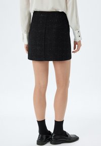 Jupe mini en tweed noir avec un motif texturé, portée avec un chemisier blanc à manches longues, des chaussettes noires et des mocassins noirs brillants.