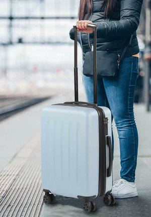 Maleta rígida de color azul claro con surcos verticales, mango negro retráctil, cuatro ruedas y cierre de cremallera, situada frente a un fondo de estación de tren.