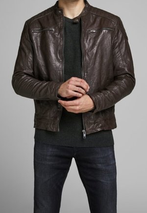 Homme portant une veste en cuir marron foncé par-dessus un pull noir et un jean bleu foncé, les mains ajustant la fermeture éclair de la veste.