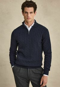 Marinblå kabelstickad tröja med zipkrage, ribbad mudd och kant. Kombinerad med grå byxor som har en klassisk och strukturerad design.