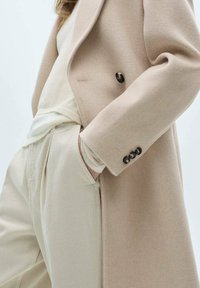 Beige ullblandad kappa med en strömlinjeformad design, med dubbelknäppt front och svarta knappdetaljer. Stylar med ljusa byxor.