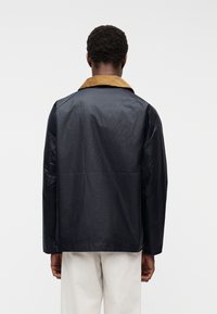 Άνδρας που φοράει σκούρο μπλε σκούρο μπουφάν με καφέ γιακά, στέκεται με την πλάτη γυρισμένη, συνδυασμένο με ανοιχτό μπεζ παντελόνι σε απλό λευκό φόντο.