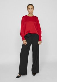Blusa rossa in raso a maniche lunghe con scollatura rotonda, abbinata a pantaloni neri a vita alta con gamba larga, completata da tacchi a punta neri.