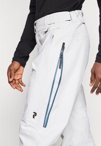 Pantalones blancos con un diseño elegante que presenta un detalle de cremallera azul y herrajes en negro. Incluye un logo identificable en el muslo.