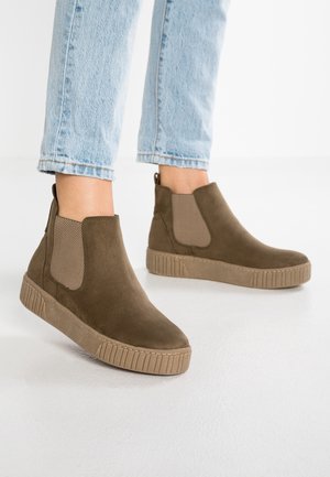 Braune Wildleder-Stiefeletten mit strukturierten elastischen Seitenpaneelen, getragen zu hellblauen, gekürzten Jeans auf weißem Boden und Hintergrund.