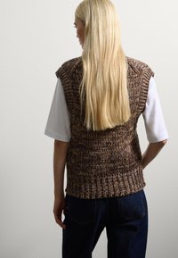 Blonde Person mit langen Haaren, die einen braunen, gestrickten, ärmellosen Pullover über einem weißen T-Shirt mit kurzen Ärmeln und dunklen Jeans trägt.