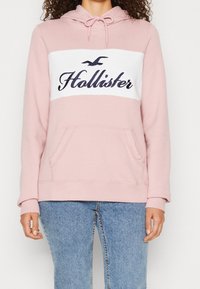 Žena v světle růžové mikině Hollister s bílým panelem a modrým logem, kombinovanou s modrými džíny, stojí na jednobarevném pozadí.