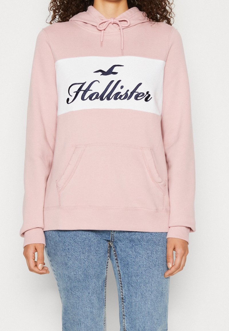 Žena v světle růžové mikině Hollister s bílým panelem a modrým logem, kombinovanou s modrými džíny, stojí na jednobarevném pozadí.